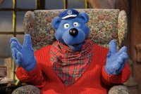 Die Puppe Käpt'n Blaubär sitzt auf einem Sessel, trägt einen roten Pullover, ein rot-kariertes Halstuch und eine blaue Schiffermütze auf dem Kopf.