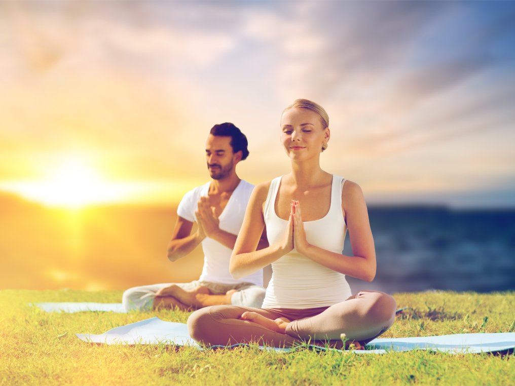 Zwei Personen sitzen im Schneidersitz auf Yogamatten im Freien und halten die Hände vor der Brust gefaltet
