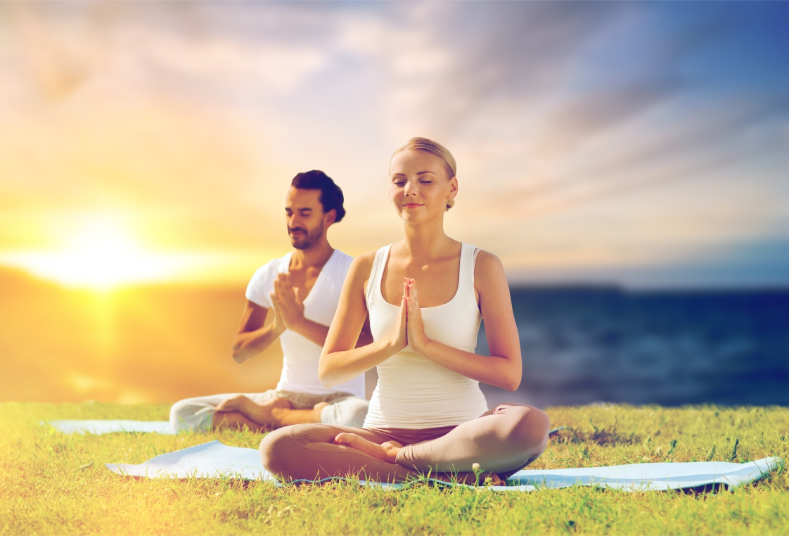 Zwei Personen sitzen im Schneidersitz auf Yogamatten im Freien und halten die Hände vor der Brust gefaltet
