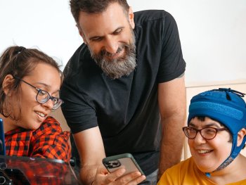 Drei lächelnde Personen schauen gemeinsam auf ein Smartphone, das eine stehende Person hält, während die beiden anderen sitzen und Brillen tragen.