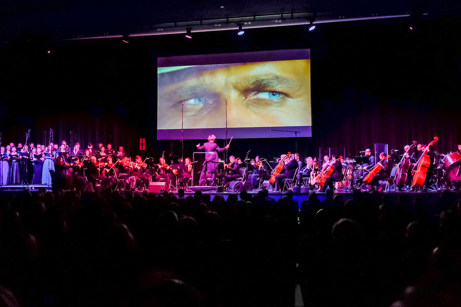 Ein großes Orchester mit Chor spielt auf einer Konzertbühne vor Publikum. Über der Bühne zeigt eine riesige Leinwand eine Nahaufnahme von blauen Augen aus einem Westernfilm – eine Anspielung auf die berühmten Filmmusiken von Ennio Morricone.