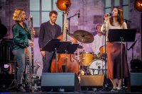 Eine Jazzband mit vier Musikern performt auf einer Bühne: Saxophonistin, Bassist, Schlagzeuger und Sängerin mit Mikrofon.