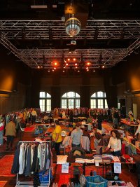 Großer Second-Hand-Kleidungsmarkt in einer Veranstaltungshalle mit rotem Teppich, zahlreichen Ständen mit Kleidungsständern und Besuchern