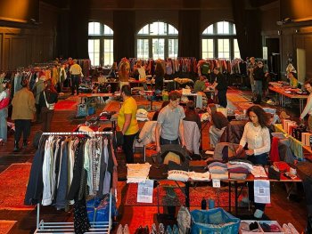 Großer Second-Hand-Kleidungsmarkt in einer Veranstaltungshalle mit rotem Teppich, zahlreichen Ständen mit Kleidungsständern und Besuchern
