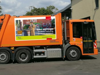 Copyright: ASN Orangefarbenes Müllfahrzeug mit Plakat "Tatort Biotonne", seitliche Ansicht auf Parkplatz.