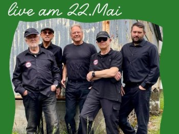 Ein Foto von fünf Personen in schwarzer Arbeitskleidung, die nebeneinander vor einer verwitterten Metallwand stehen. Oben links steht in weißer Schreibschrift „live am 22.Mai".