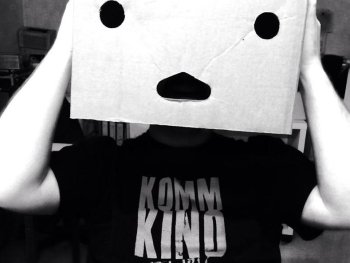 Copyright: Kommkino e. V. Person mit weißem Papier als Kopfbedeckung, auf dem ein überraschtes Gesicht gezeichnet ist, trägt schwarzes Shirt mit "KOMM KIND"-Schriftzug.