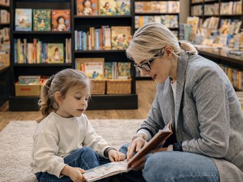 Eine erwachsene Person und ein Kind sitzen auf einem Teppich und lesen gemeinsam ein Buch, umgeben von vollen Bücherregalen.