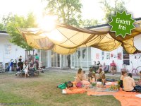 Menschen sitzen und liegen auf orangefarbenen Decken unter einem großen, gelb-braunen Tuchdach im Hof vor einem weißen Gebäude mit Glastüren. An den Wänden hängen Poster, daneben stehen Fahrräder. Die Szene ist hell und sonnig.