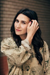 Portrait Yade Önder: Person mit dunklem, welligem Haar posiert nachdenklich mit der Hand am Kopf vor einer Backsteinwand.
