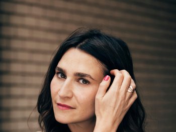 Portrait Yade Önder: Person mit dunklem, welligem Haar posiert nachdenklich mit der Hand am Kopf vor einer Backsteinwand.