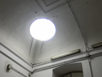 Foto: Nicolas Hansmann
Rundes Oberlicht in einer hohen, weißen Raumdecke mit sichtbaren Kabeln und Leuchtstoffröhre an der Wand