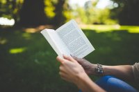 Person hält aufgeschlagenes Buch in beiden Händen im Freien sitzend