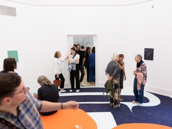 Copyright: Cornelia Baltes, Kir, 2026 Besucherinnen und Besucher unterhalten sich in einem Ausstellungsraum mit weißen Wänden, kleinen Kunstwerken und einem auffällig gemusterten Boden.