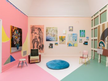 Foto: Lukas Pürmayr
Bunt gestalteter Ausstellungsraum mit rosa und grün bemalten Wänden, zahlreichen Gemälden unterschiedlicher Größe und Stile sowie einem Sessel, Stuhl, Hocker und einem runden blauen Kissen auf dem Boden.