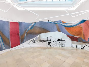 Foto: © VG Bild-Kunst Bonn, 2026 für Henriette Grahnert Foto: Lukas Pürmayr
Ein großformatiger, wellenförmig von der Decke hängendes bedruckter Teppich mit abstrakter Malerei in Blau, Orange und Rot hängt im Raum, der an den Wänden mit schwarzen Linienzeichnungen bemalt ist.