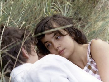 Copyright: Les Films du Losange Zwei Personen liegen entspannt im hohen Gras, wobei eine Person mit dunklem Haar und kariertem Top in die Kamera blickt.