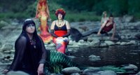 Mehrere als Merfolk verkleidete Personen mit bunten Kostümen und Flossen sitzen auf Steinen an einem Fluss in einer Waldlandschaft.