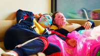 Zwei Personen mit bunten Gesichtsmasken liegen entspannt auf einer Couch, umgeben von pinken Folienstoffen.