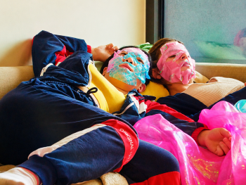 Copyright: Excellent Friends Zwei Personen mit bunten Gesichtsmasken liegen entspannt auf einer Couch, umgeben von pinken Folienstoffen.