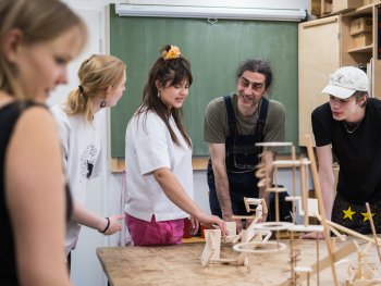 Eine lehrende Person erklärt mehreren Jugendlichen in einem Werkraum verschiedene Holzkonstruktionen, die auf einem Werktisch stehen.