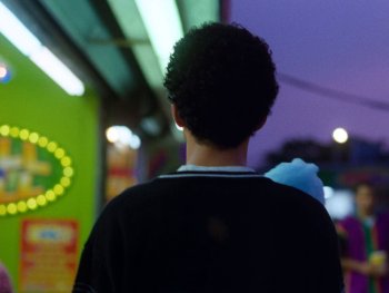 Copyright: A24 Person von hinten auf einer abendlichen Marktstraße mit bunten Neonlichtern und lila Abendhimmel.