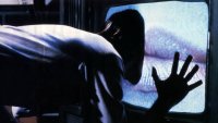 Eine Person beugt sich in einer dunklen Szene mit einer Hand an einen alten Röhrenfernseher, dessen Bildschirm statisches Rauschen zeigt.