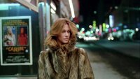 Eine Person in einem Pelzmantel steht nachts auf einem Bürgersteig vor einem Kinoplakat des Films „Carrie" (1976).