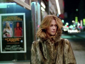 Copyright: Filmverleih Eine Person in einem Pelzmantel steht nachts auf einem Bürgersteig vor einem Kinoplakat des Films „Carrie" (1976).