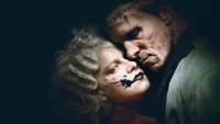 Zwei Personen mit theatralisch-dunklem Make-up liegen Wange an Wange in einer düsteren, dramatischen Szene.