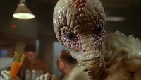 Detailaufnahme einer aufwendigen Alien-Figur mit faltiger, feuchter Haut und großem Schädel, die einen orangefarbenen Drink in einer Bartheke hält.