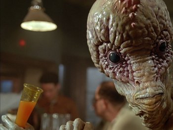 Copyright: Filmverleih
Detailaufnahme einer aufwendigen Alien-Figur mit faltiger, feuchter Haut und großem Schädel, die einen orangefarbenen Drink in einer Bartheke hält.