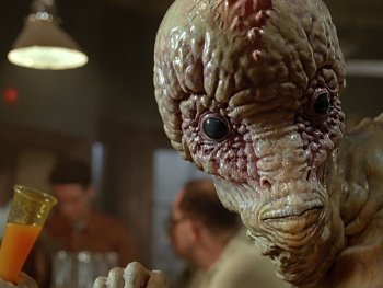 Copyright: Filmverleih Detailaufnahme einer aufwendigen Alien-Figur mit faltiger, feuchter Haut und großem Schädel, die einen orangefarbenen Drink in einer Bartheke hält.