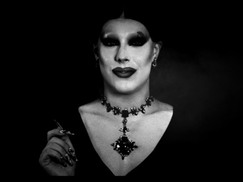 Copyright: Filmverleih Person in aufwendigem Gothic-Drag-Make-up mit schwarzem Augen-Make-up, Schmuckkette und langen Nägeln in schwarz-weißer Porträtaufnahme.