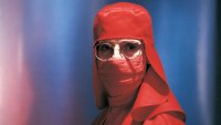 Person in einem vollständig roten Schutzanzug mit Haube, Gesichtsmaske und Brille blickt frontal in die Kamera.