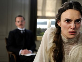 Copyright: Filmverleih
Eine junge Person in historischer weißer Kleidung blickt angespannt in die Kamera, im unscharfen Hintergrund sitzt eine weitere Person in dunklem Anzug.