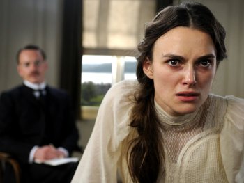 Copyright: Filmverleih Eine junge Person in historischer weißer Kleidung blickt angespannt in die Kamera, im unscharfen Hintergrund sitzt eine weitere Person in dunklem Anzug.