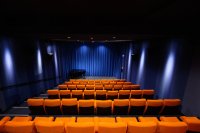 Der Saall kommkino vom Filmhaus: leerer Saal mit orangefarbenen Sitzreihen, blau beleuchtetem Bühnenvorhang und einem schwarzen Flügel auf der Bühne.