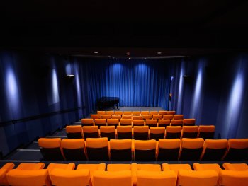 Der Saall kommkino vom Filmhaus: leerer Saal mit orangefarbenen Sitzreihen, blau beleuchtetem Bühnenvorhang und einem schwarzen Flügel auf der Bühne.