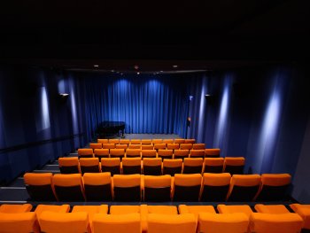 Der Saall kommkino vom Filmhaus: leerer Saal mit orangefarbenen Sitzreihen, blau beleuchtetem Bühnenvorhang und einem schwarzen Flügel auf der Bühne.