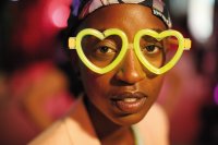 Eine Person trägt eine neongelbe Herzform-Brille und ein gemustertes Kopftuch und schaut direkt in die Kamera.