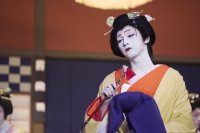 Person in traditioneller japanischer Kabuki-Kostümierung mit weißer Gesichtsschminke, gelbem Kimono und aufwendiger Kopfbedeckung hält einen Fächer.