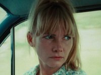 Eine blonde Person mit Ponyfransen sitzt in einem Auto und blickt mit ernstem Gesichtsausdruck zur Seite.