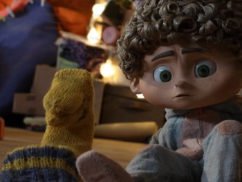 Copyright: Filmverleih
Animierte Kinderfigur mit lockigem Haar und großen grünen Augen sitzt nachdenklich zwischen Wollsocken in einer warm beleuchteten Umgebung.
