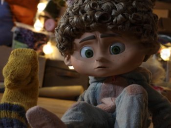 Copyright: Filmverleih Animierte Kinderfigur mit lockigem Haar und großen grünen Augen sitzt nachdenklich zwischen Wollsocken in einer warm beleuchteten Umgebung.