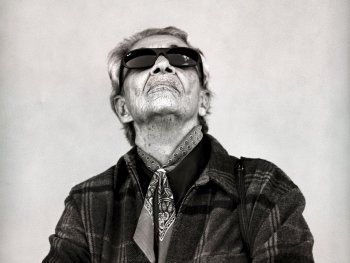 Chavela mit Sonnenbrille und karierter Jacke, Arme verschränkt, Blick nach oben gerichtet.