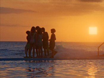 Copyright: The Barricades; Panama Film; Gregory Oke Silhouetten mehrerer Personen stehen bei Sonnenuntergang am Rand eines Außenpools mit Meerblick im goldenen Abendlicht.