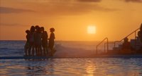 Silhouetten mehrerer Personen stehen bei Sonnenuntergang am Rand eines Außenpools mit Meerblick im goldenen Abendlicht.