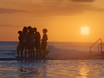 Copyright: The Barricades; Panama Film; Gregory Oke Eine Gruppe von Personen steht am Rand eines Meerwasserpools und blickt bei Sonnenuntergang auf das Meer.