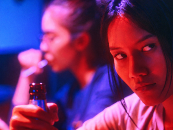 Copyright: 2026 eksystent Filmverleih
Nahaufnahme einer jungen Frau in rot-blauem Neonlicht, die mit ernstem Blick zur Seite schaut, im Hintergrund eine Person mit einer Getränkeflasche.
